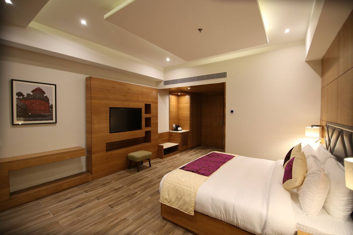 A modern hotel room with a large bed, a flat-screen TV mounted on the wall, a wooden bench, and a small table.