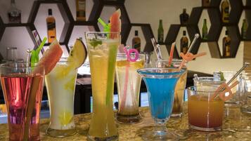 Diversos cocktails coloridos sobre um balcão de bar, com um expositor de garrafas hexagonal ao fundo.