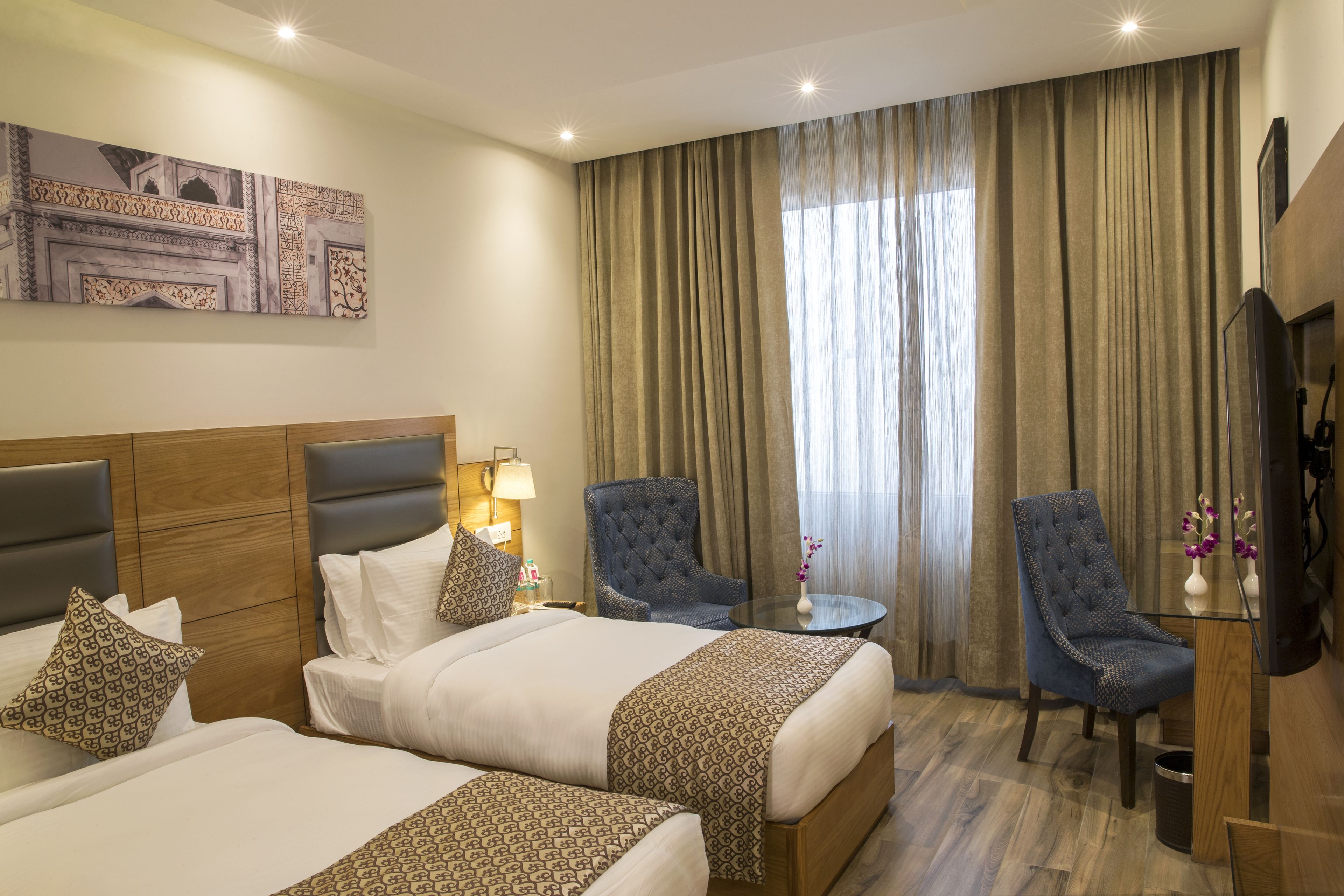 A hotel room with two beds, a desk, a chair, a TV, and a large window with curtains.