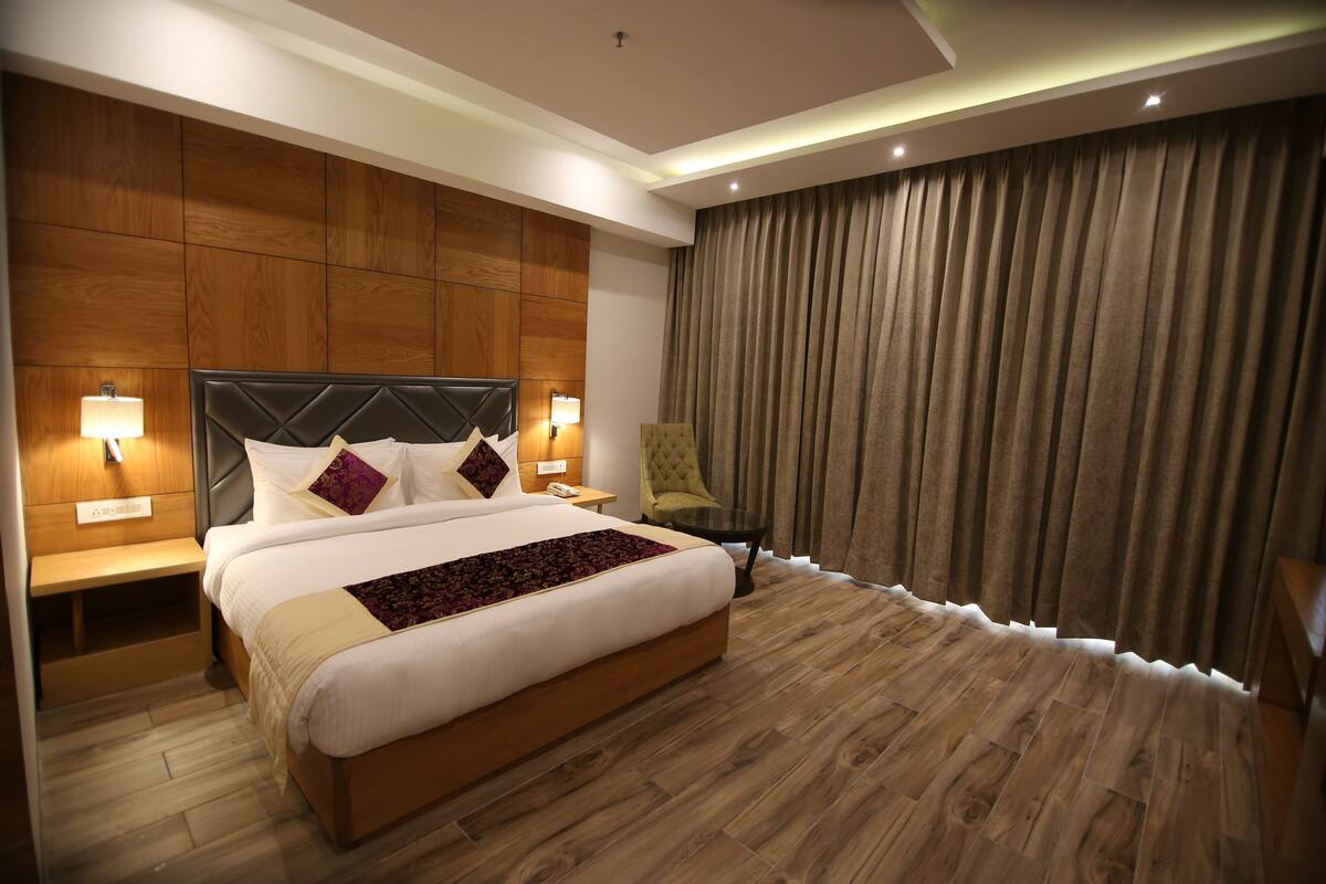 A modern hotel room with a large bed, wooden paneling on the walls, a large window with curtains, and a small table with a lamp.