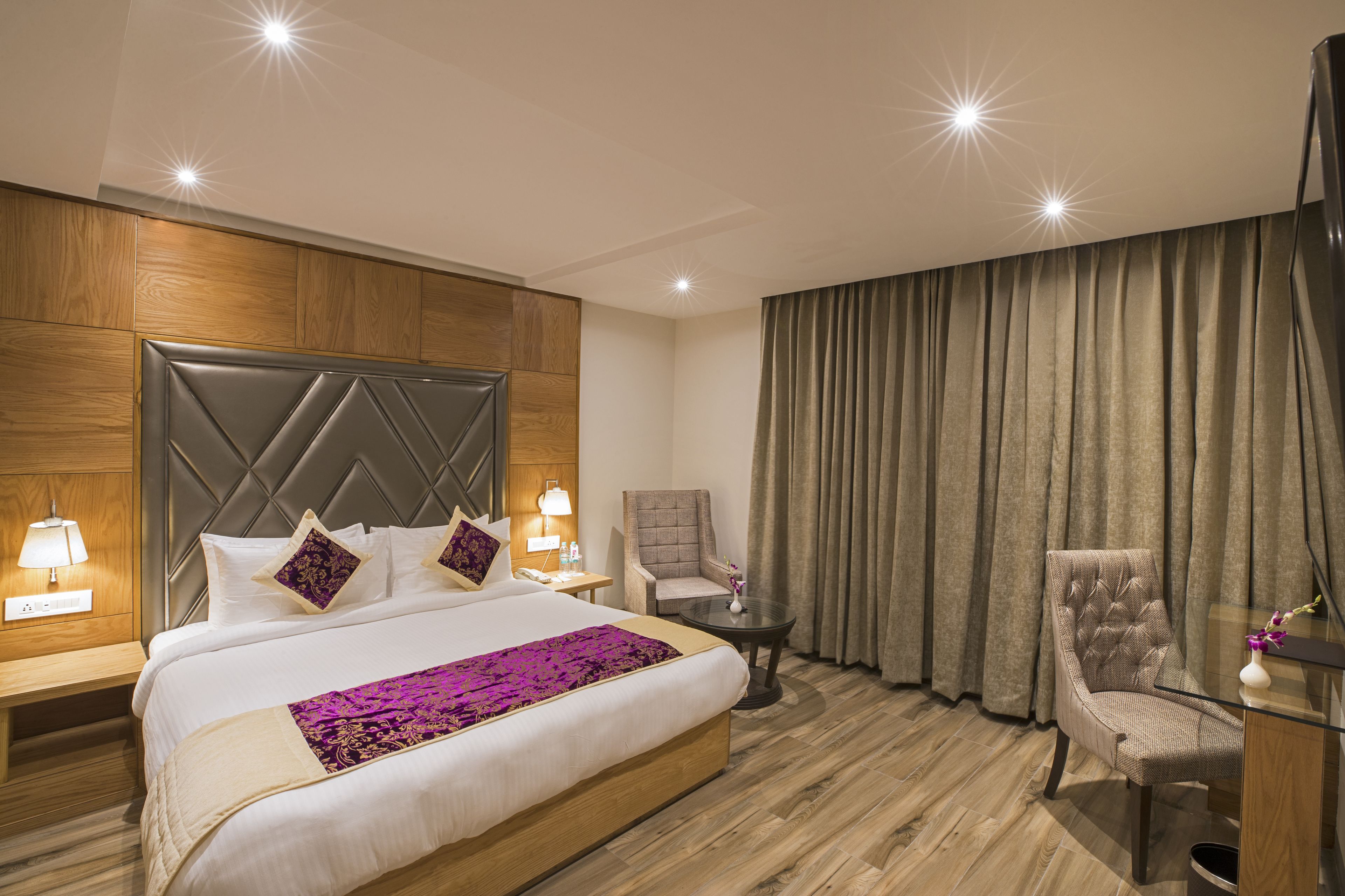 A hotel room with a large bed, two armchairs, a small table, and a nightstand with a lamp.