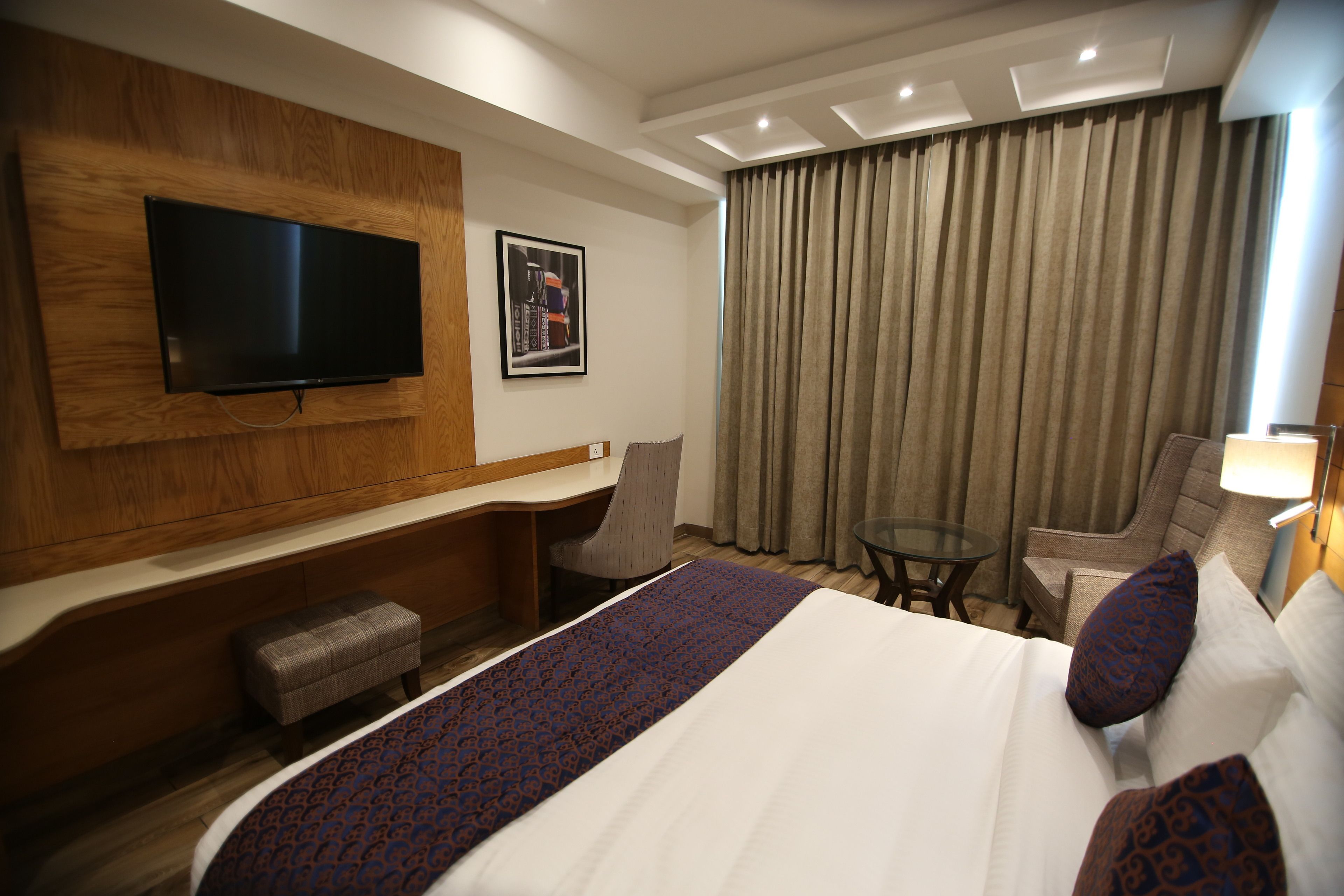 A hotel room with a large bed, a flat-screen TV mounted on a wooden panel, a desk with a chair, a small round table, and a large window with curtains.