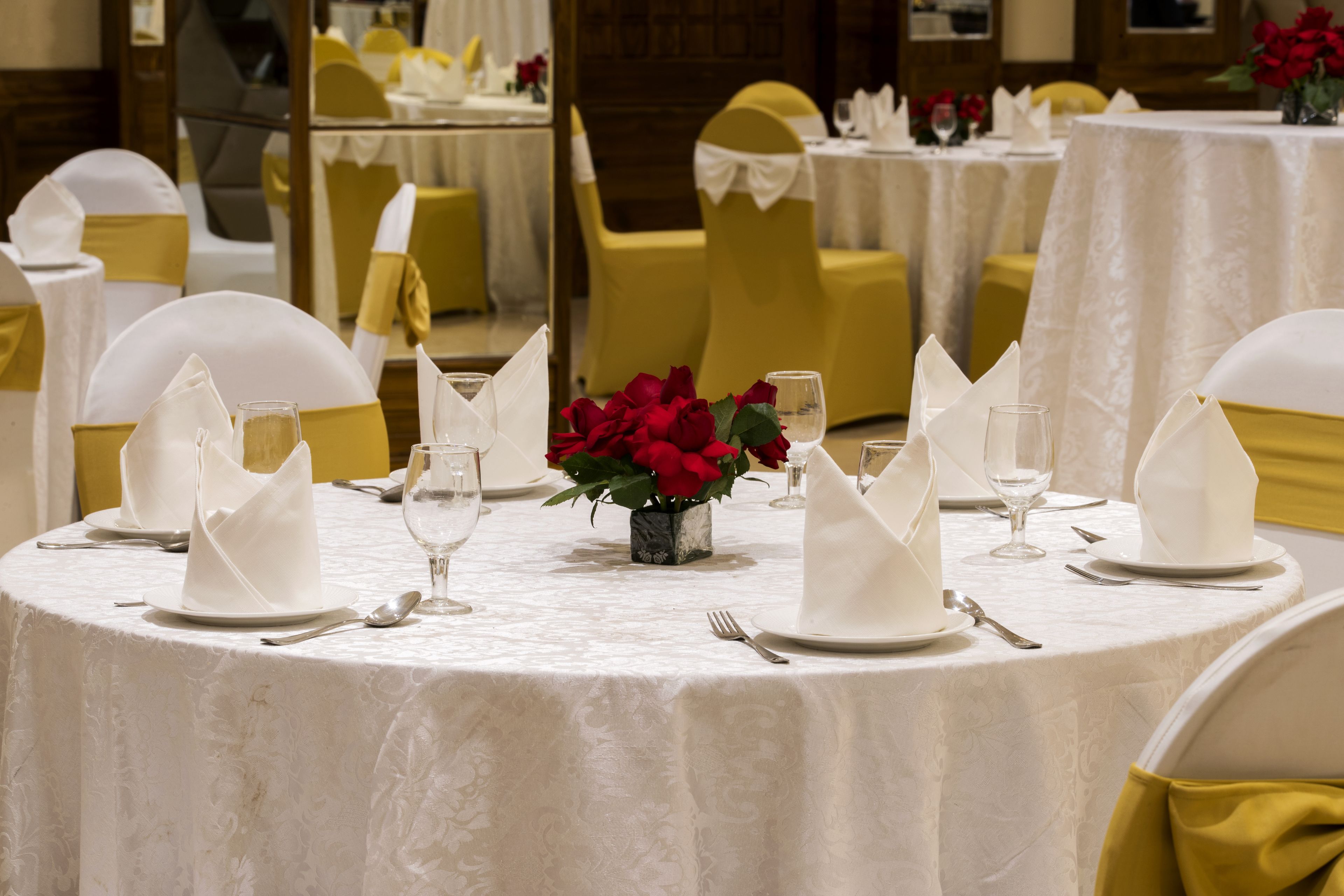 Un salón de banquetes con mesas redondas dispuestas para un evento formal, con manteles blancos, fajas doradas y centros de mesa con rosas rojas.