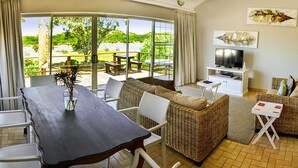 A living room with a dining table, wicker sofa, and a TV on a white stand. - Addo River-View Lodge (Colchester)