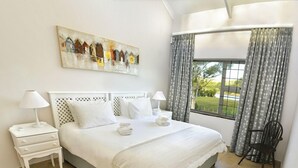A neatly arranged hotel room with a large bed, two bedside tables, a chair, a window with curtains, and a painting on the wall. - Addo River-View Lodge (Colchester)