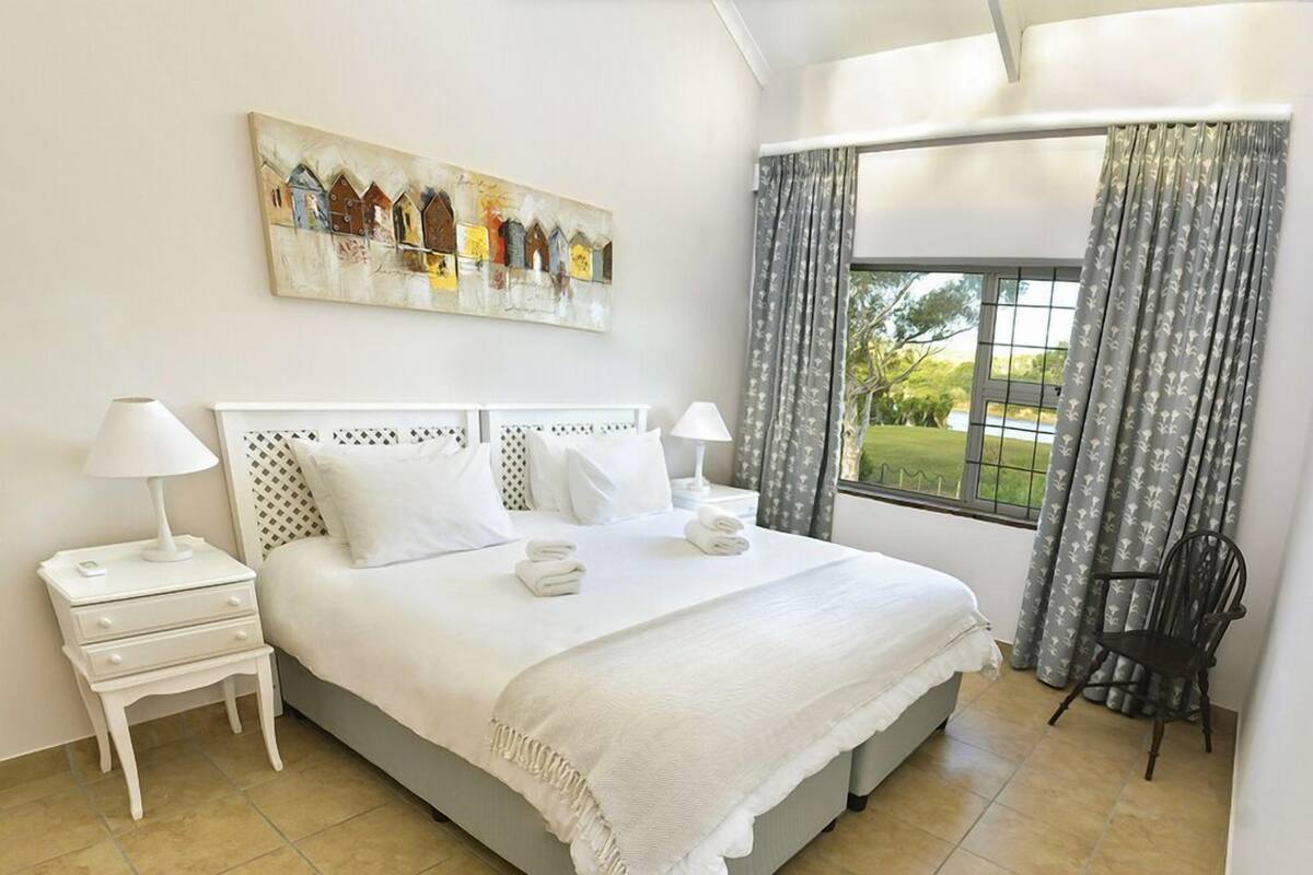 A neatly arranged hotel room with a large bed, two bedside tables, a chair, a window with curtains, and a painting on the wall.