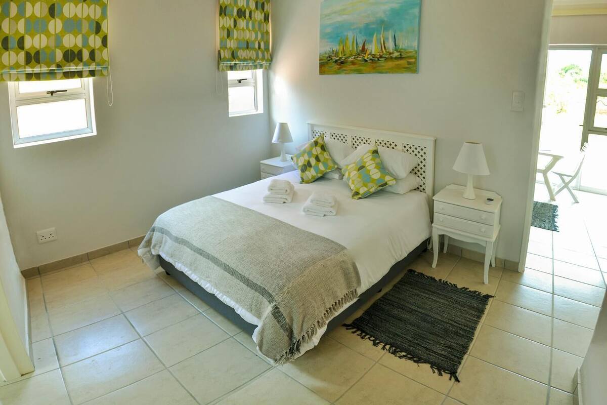 A bedroom with a bed, two bedside tables, a painting, and a window with blinds.