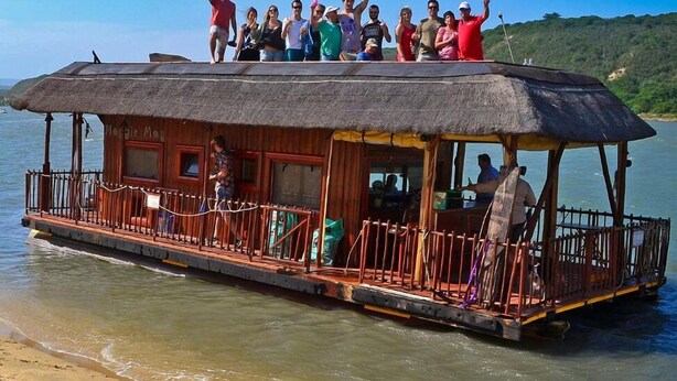 A boat with a thatched roof and wooden deck, passengers on the upper deck, and a sandy beach in the background. - Addo River-View Lodge (Colchester)