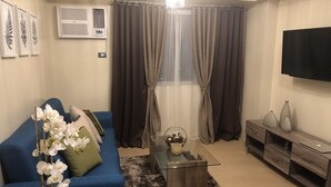 Una sala de estar con un sofá azul, una mesa de centro de vidrio, un televisor montado en la pared y un mueble de madera.