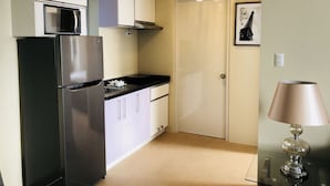 Una cocina compacta con refrigerador, microondas y una pequeña encimera. Hay una puerta que da a otra habitación y una lámpara sobre una mesa.