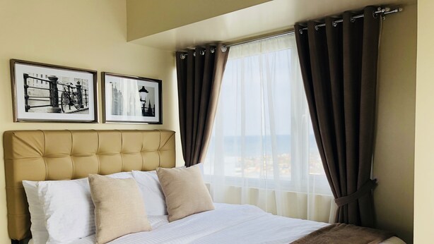 Una cama bien hecha con cabecera, dos cuadros enmarcados en la pared y vista al mar a través de cortinas translúcidas.