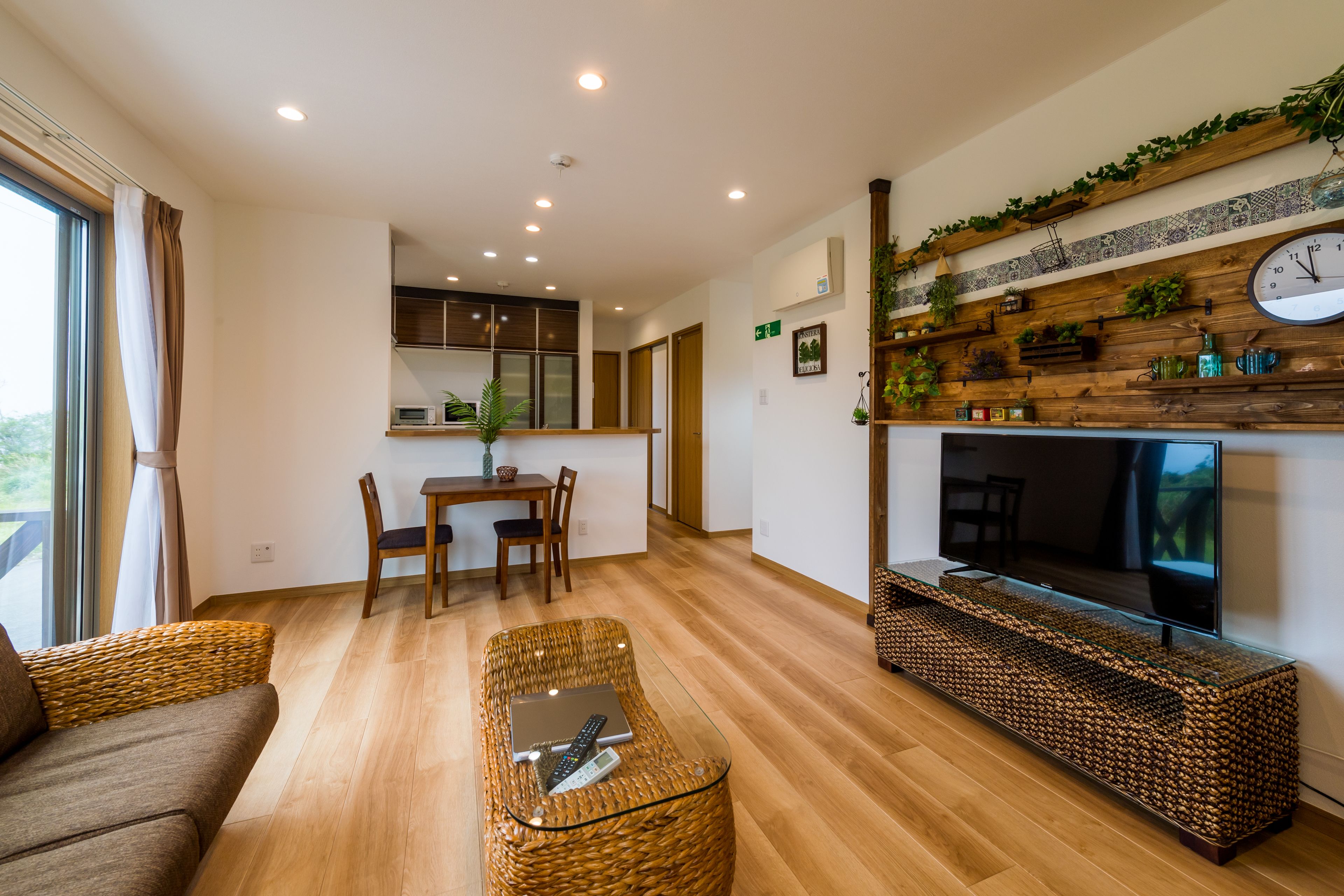 A modern living room with a sofa, a glass coffee table with a laptop and remote, a TV on a wooden shelf, and a dining area with a table and chairs.