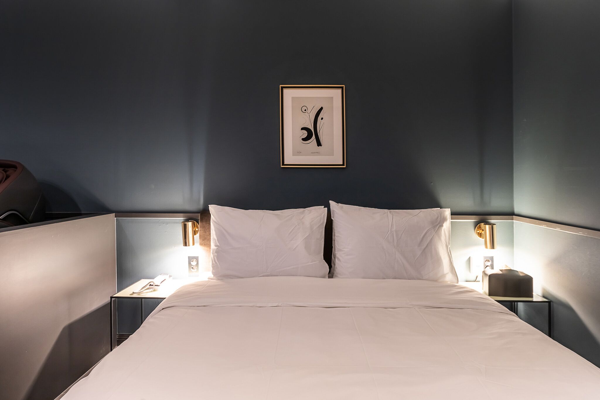 Quarto de hotel com uma cama grande, duas mesas de cabeceira, uma obra de arte emoldurada na parede e uma mesa de apoio com uma luminária.