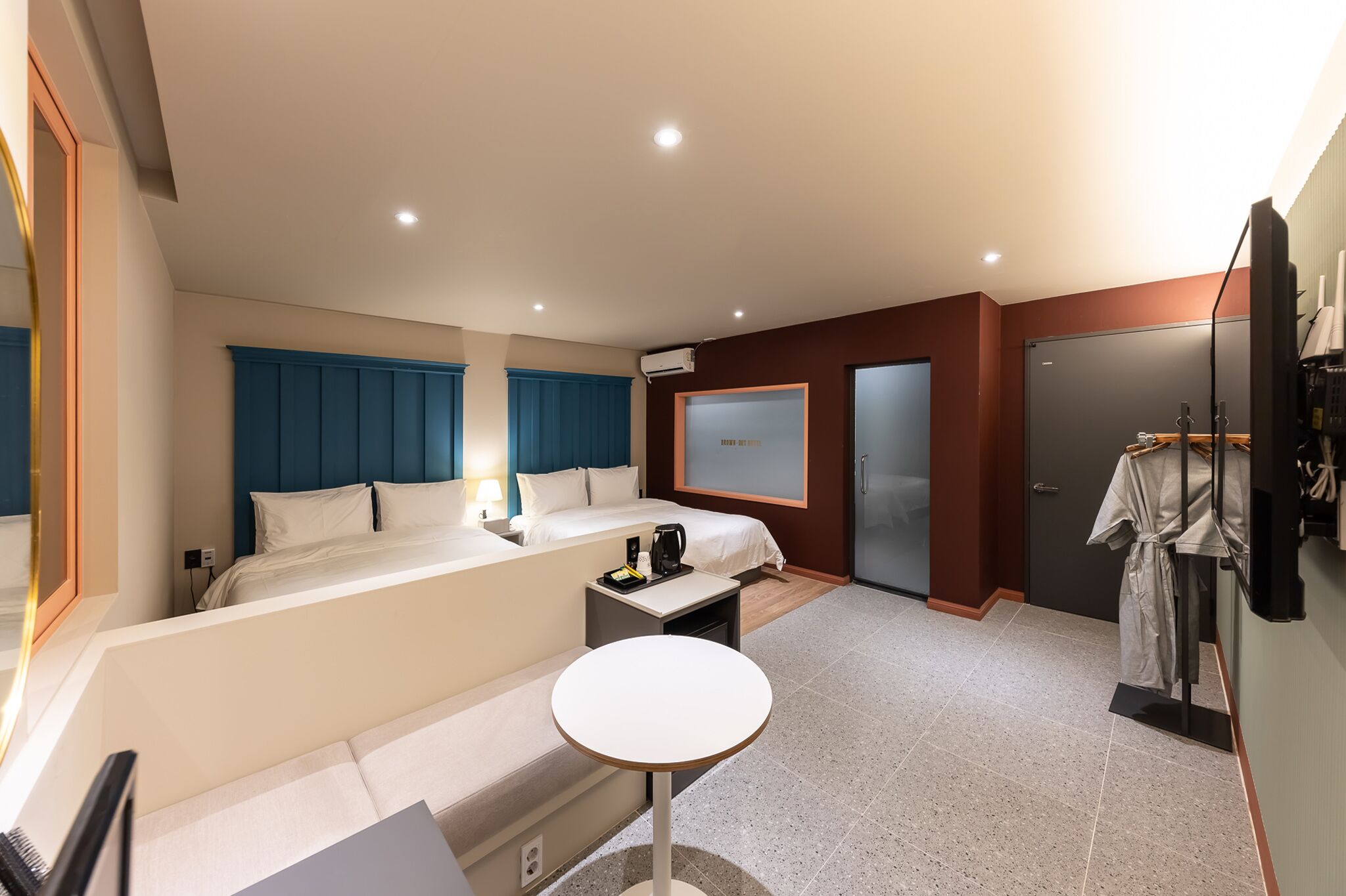 A hotel room with a bed, bedside table, and a small round table. There is a wardrobe, a mirror, and a TV on the wall.