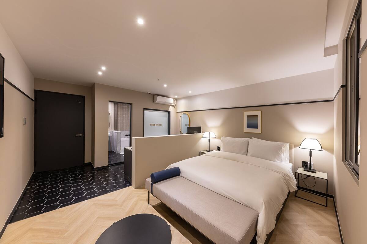 A modern hotel room with a large bed, bedside tables with lamps, a small desk, and a bathroom visible through an open door.