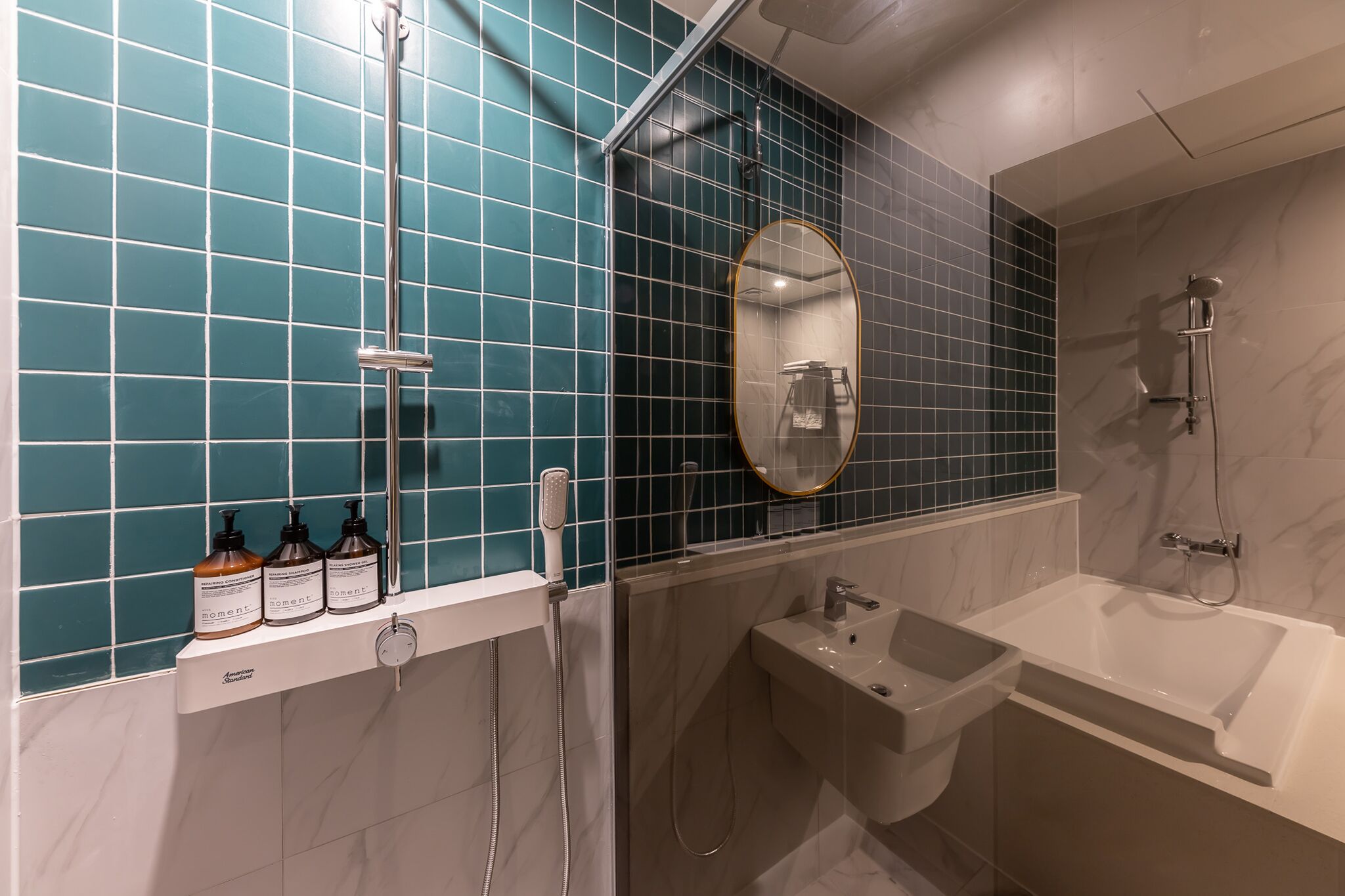 A modern bathroom with teal tiles, a large mirror, a bathtub, a shower, and three bottles of shampoo.