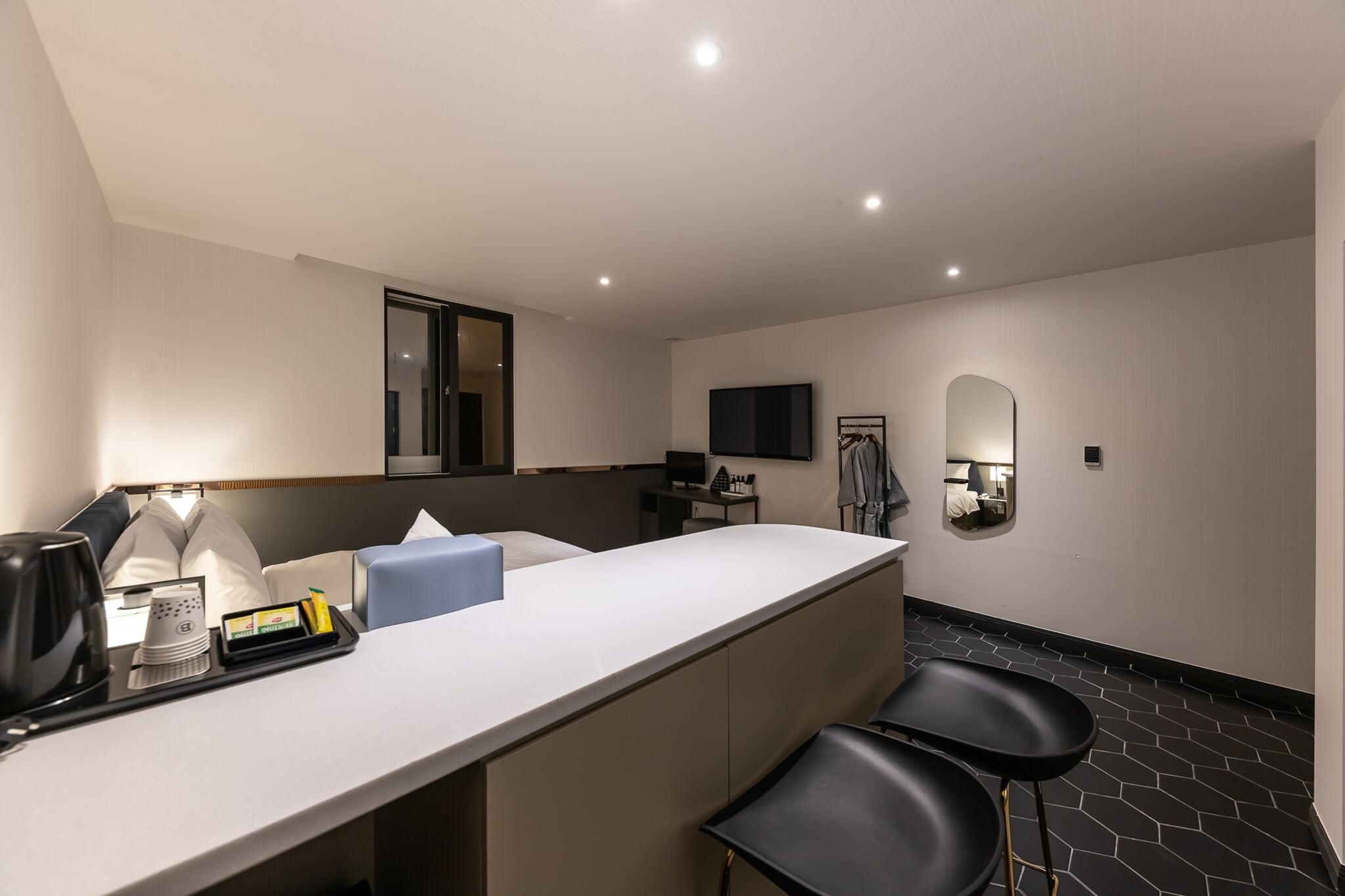 Una habitación de hotel moderna con un mostrador de recepción blanco, sillas negras, un televisor de pantalla plana, un espejo y una mesita de noche con artículos de aseo.