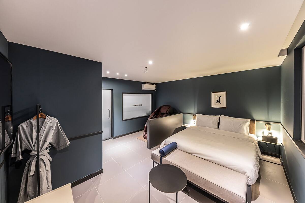 A modern hotel room with a large bed, a bedside table with a lamp, a robe hanging on a rack, and a large window.