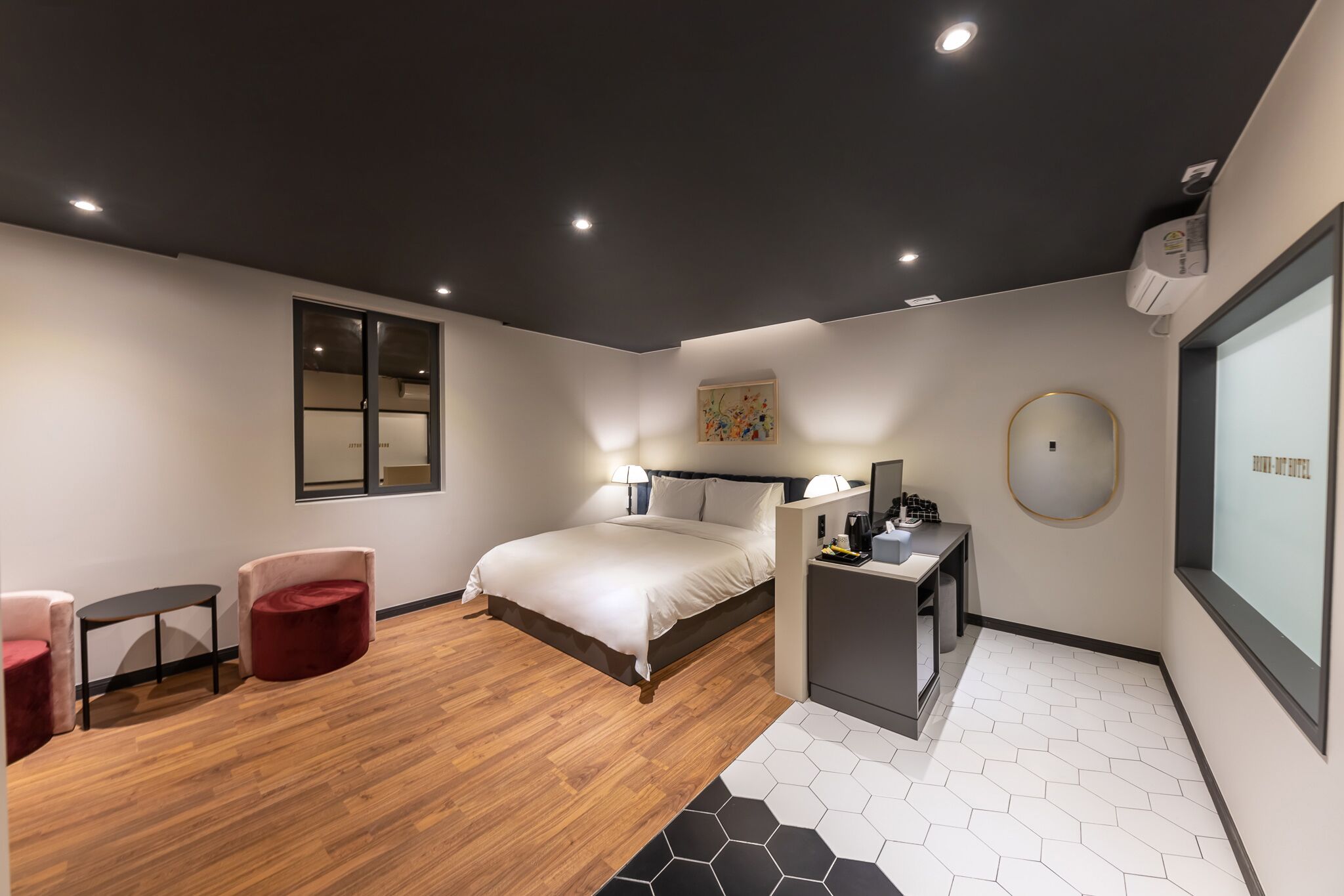 A modern hotel room with a bed, two chairs, a small table, a desk, and a large window.