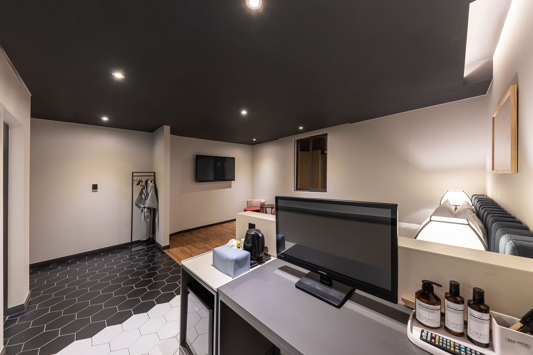 Habitación de hotel moderna con televisión de pantalla plana, escritorio con ordenador y zona de estar con sofá y lámparas.