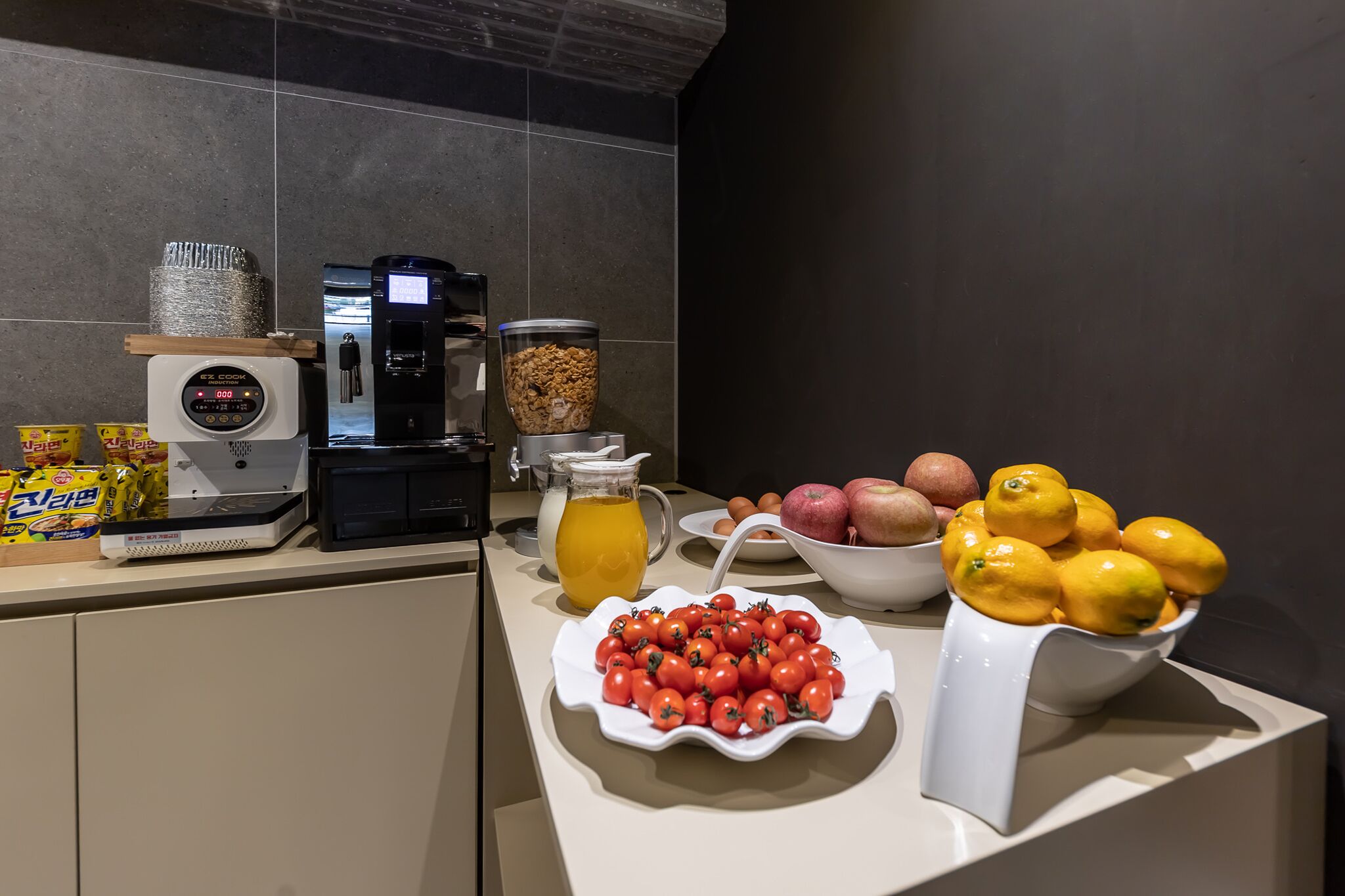 Una encimera de cocina con una cafetera, un exprimidor de frutas, un tazón de tomates, un tazón de limones y una jarra de jugo de naranja.