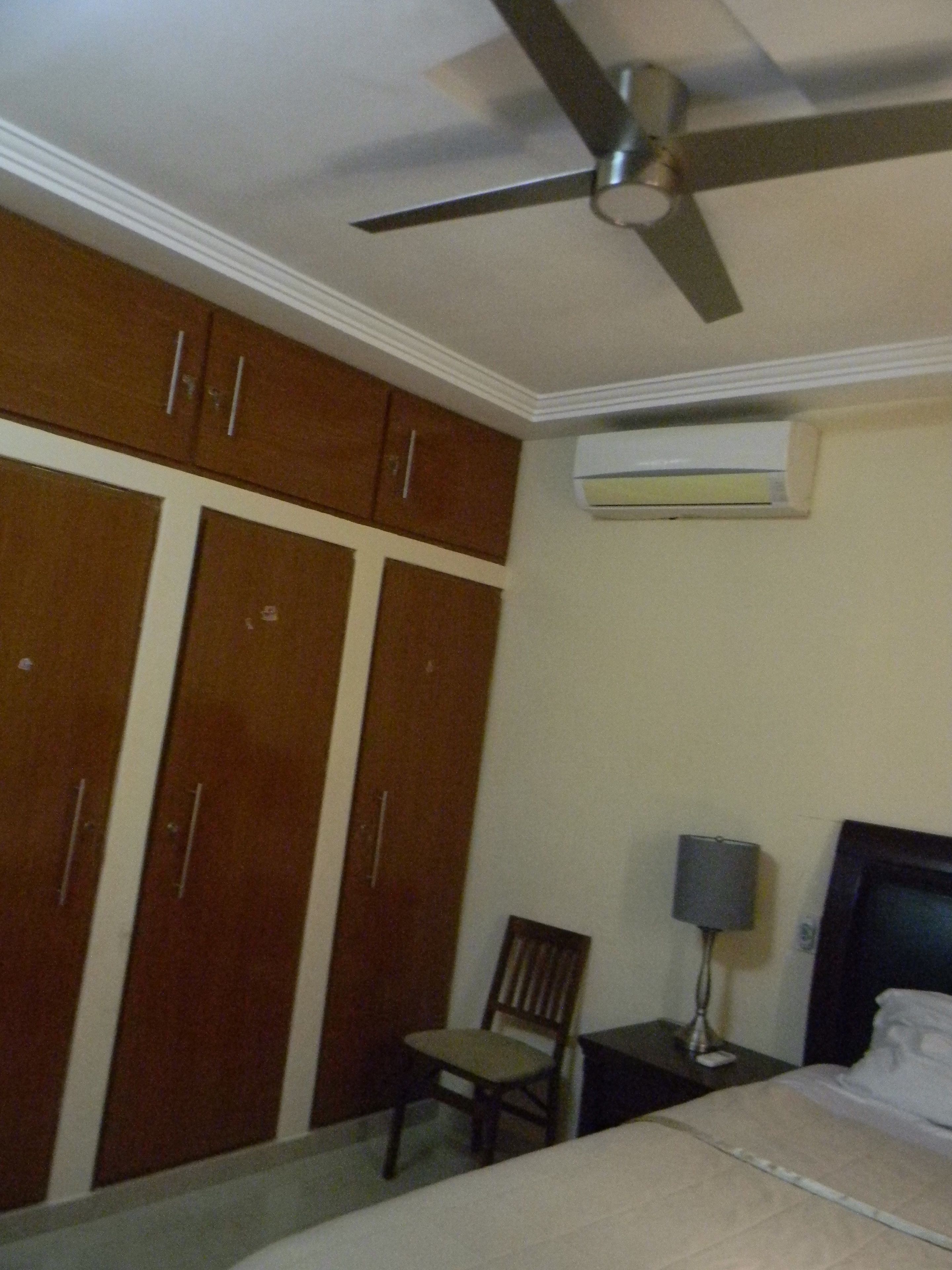 A bedroom with a bed, a chair, a desk with a lamp, a ceiling fan, and a wardrobe.