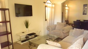 Een woonkamer met een bank, een glazen salontafel, een aan de muur bevestigde televisie en een plant op een plank.