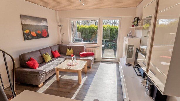 Ein Wohnzimmer mit Sofa, Couchtisch und Fernseher. Ein Fenster bietet Blick auf grüne Natur.