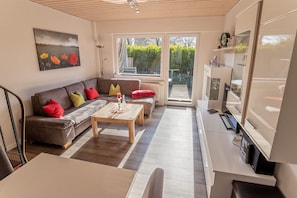 Ein Wohnzimmer mit Sofa, Couchtisch und Fernseher. Ein Fenster bietet Blick auf grüne Natur.