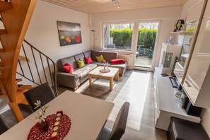A modern living room with a sofa, coffee table, and a dining table. There is a staircase leading to another level, and a view of a garden through large windows.