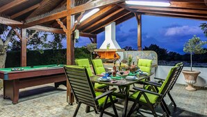 Een terras buiten met een pooltafel, groene fauteuils met kussens en een stenen open haard.