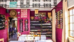 A wine shop with a variety of wine bottles displayed on shelves and walls, a sign that reads "It's all about the wine," and a table set for dining. - Sani Mountain Lodge (Sani Pass)
