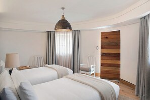 A hotel room with two beds, a lamp, a chair, a wooden door, and a hanging light fixture. - Sani Mountain Lodge (Sani Pass)