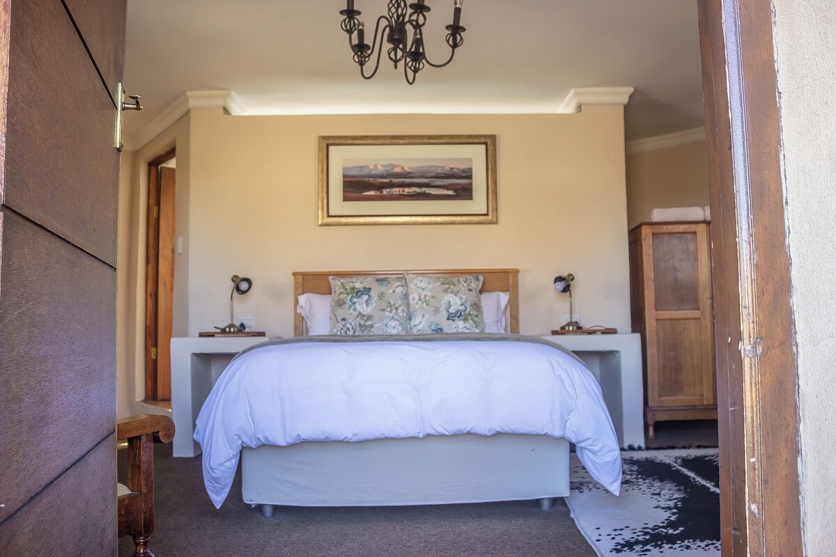 A bedroom with a large bed, a wooden headboard, a bedside table, a nightstand, a chandelier, a framed picture on the wall, and a wooden wardrobe.