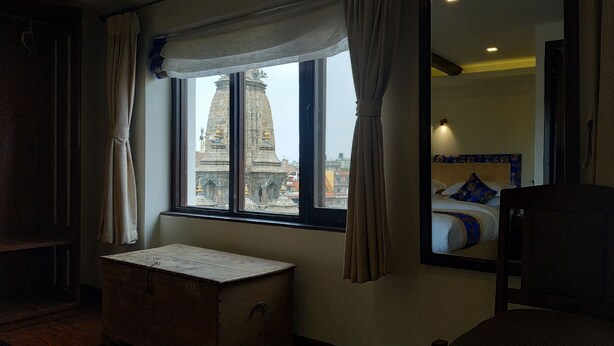 Habitación de hotel con un ventanal grande, un cofre de madera, una cama con ropa de cama azul y blanca, y un espejo que refleja la cama.