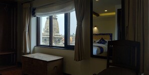 Habitación de hotel con un ventanal grande, un cofre de madera, una cama con ropa de cama azul y blanca, y un espejo que refleja la cama.