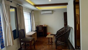 Una habitación pequeña y acogedora con piso de madera, un escritorio con silla, un sillón de mimbre, una ventana con cortinas y un equipo de aire acondicionado en la pared.