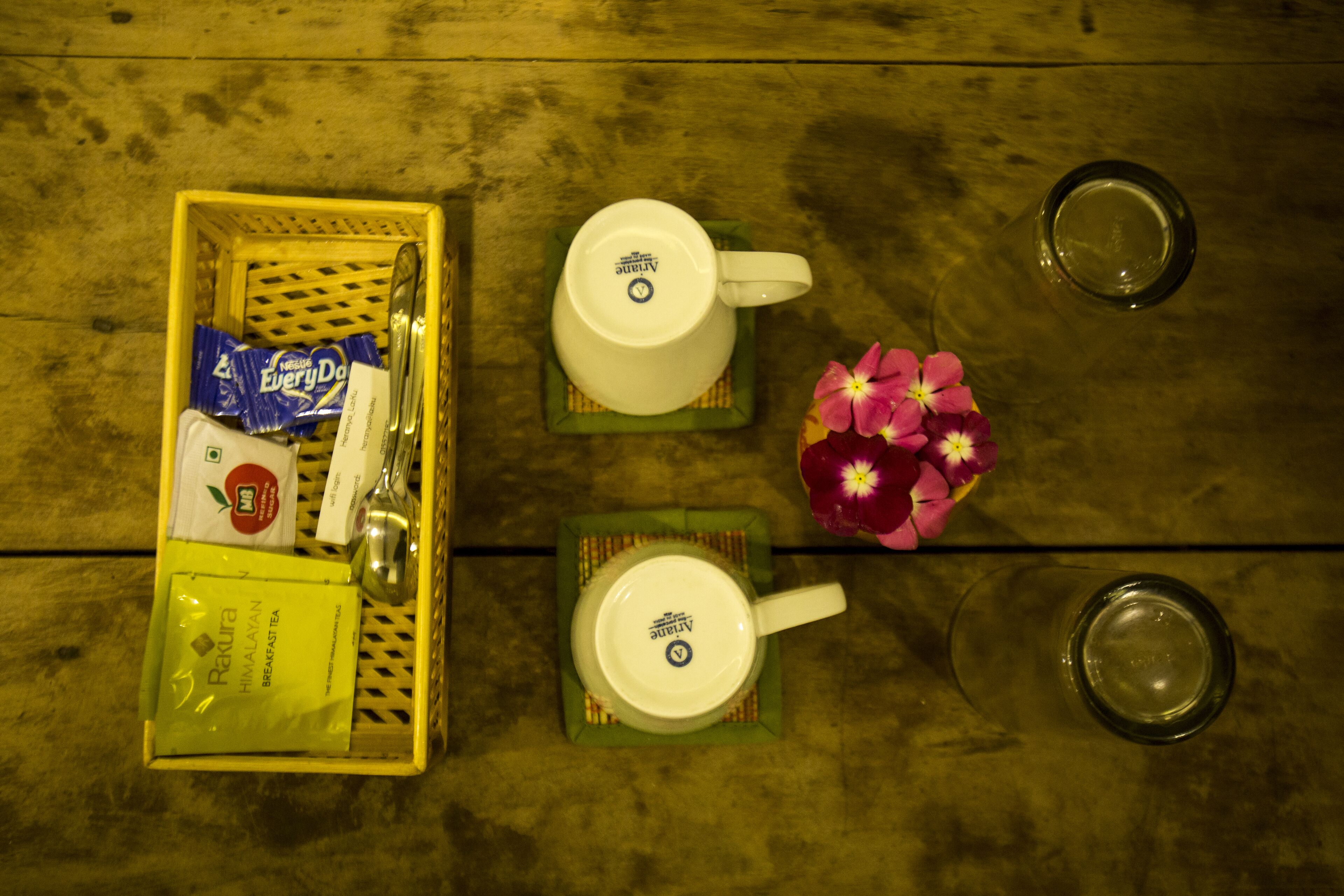 A wooden table with a tray containing a white mug, a glass of water, a packet of tea, and a small vase with pink flowers.
