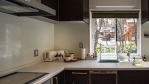 Una cocina moderna con estufa integrada, armarios oscuros, un ventanal con vistas a edificios y una encimera con diversos electrodomésticos y objetos.