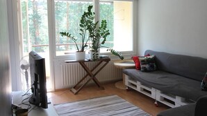 En moderne stue med en grå sofa, et lille hvidt bord, en potteplante og et stort vindue med udsigt til træer.