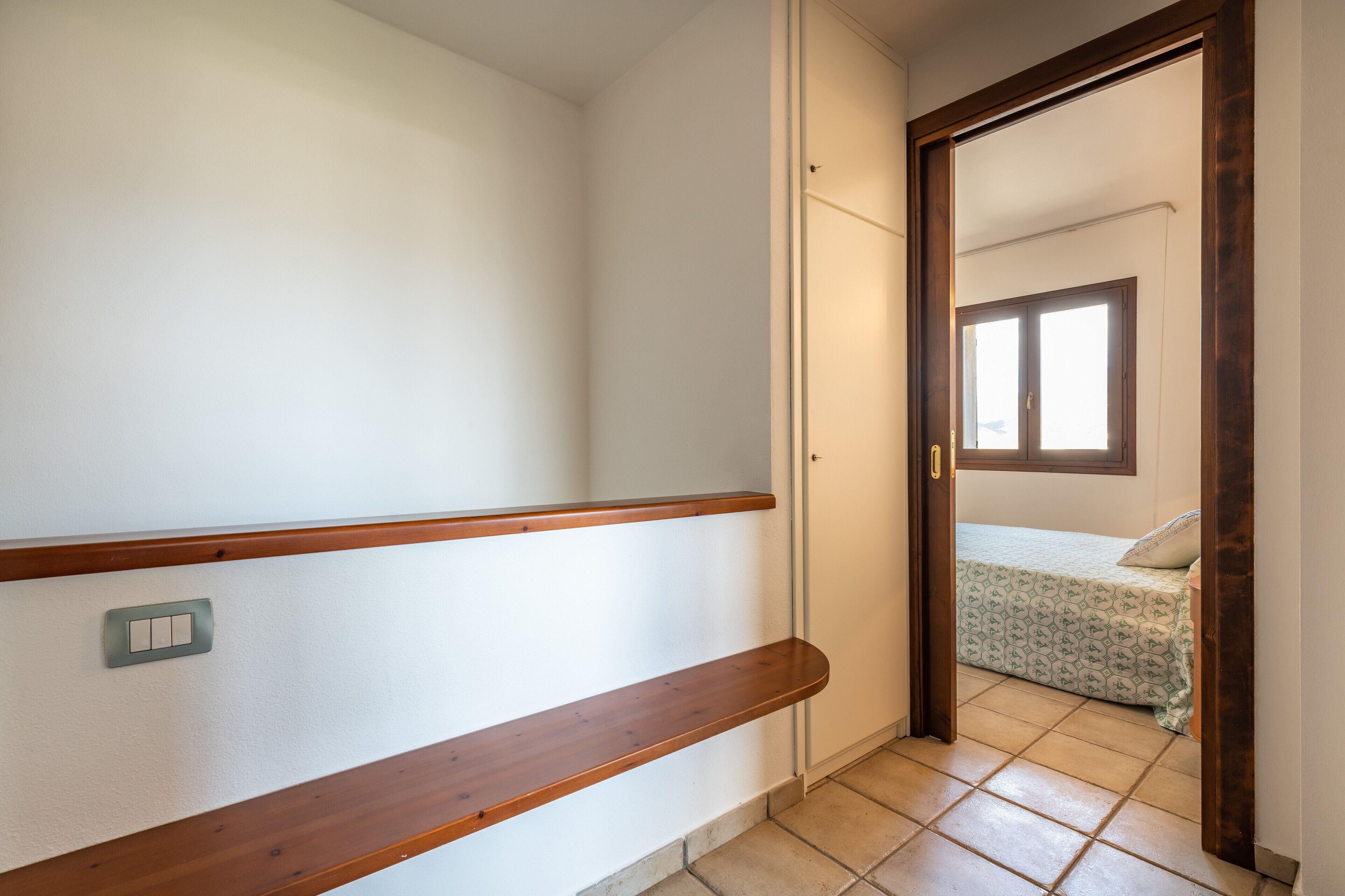 A hallway with a wooden handrail, a light switch, and a door leading to a room with a bed and window.