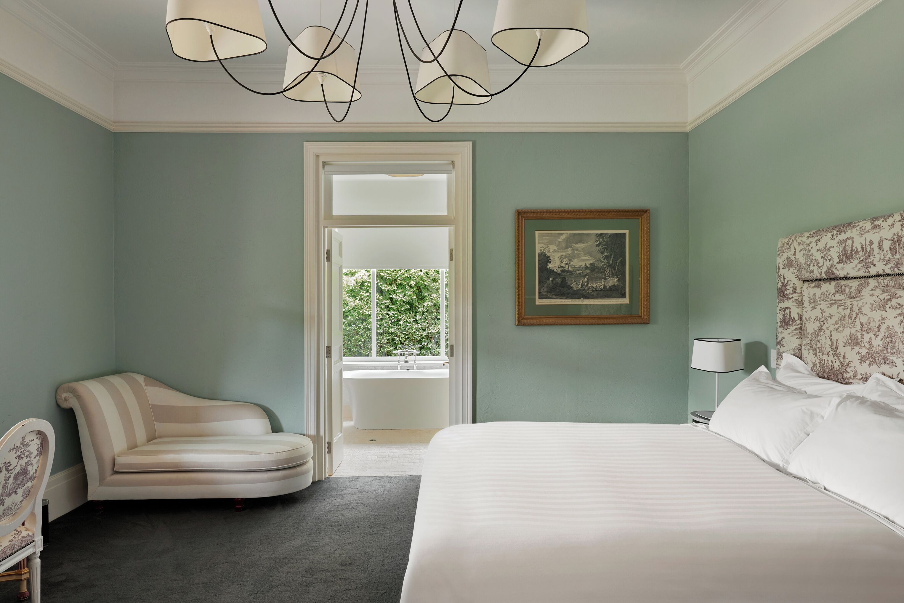 A bedroom with a bed, a chair, a painting, a lamp, and a view of a window and a bathtub.