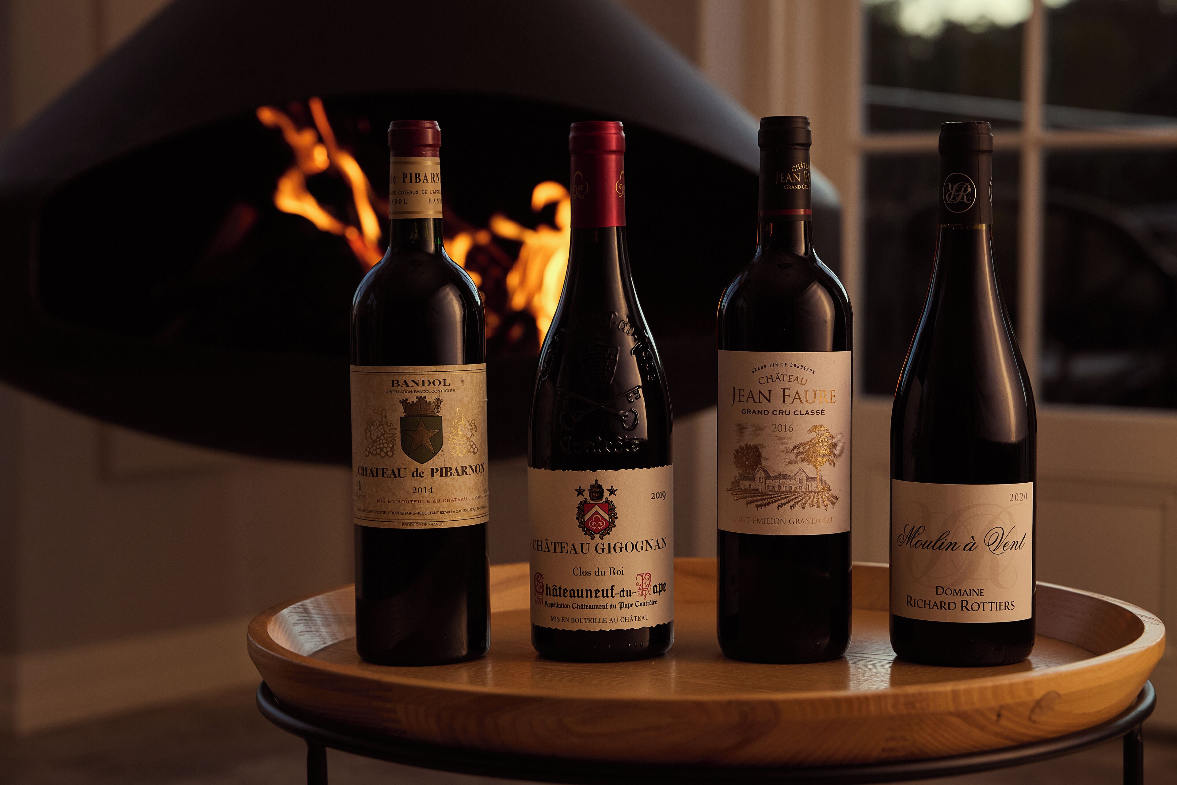 Quatre bouteilles de vin sur un plateau en bois, devant une cheminée.