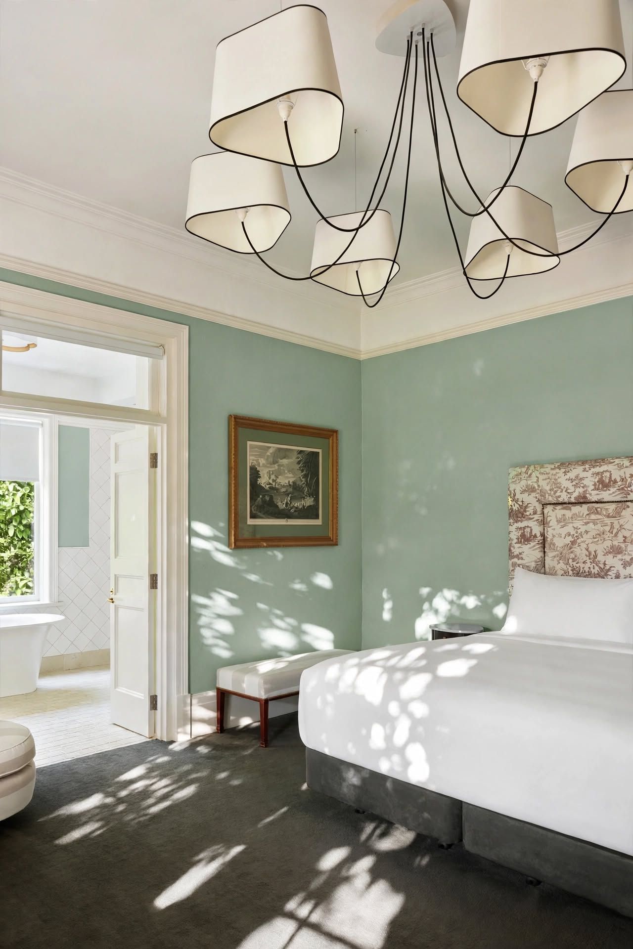 A bedroom with a bed, a bedside table, a framed picture on the wall, and a window with a view of greenery.