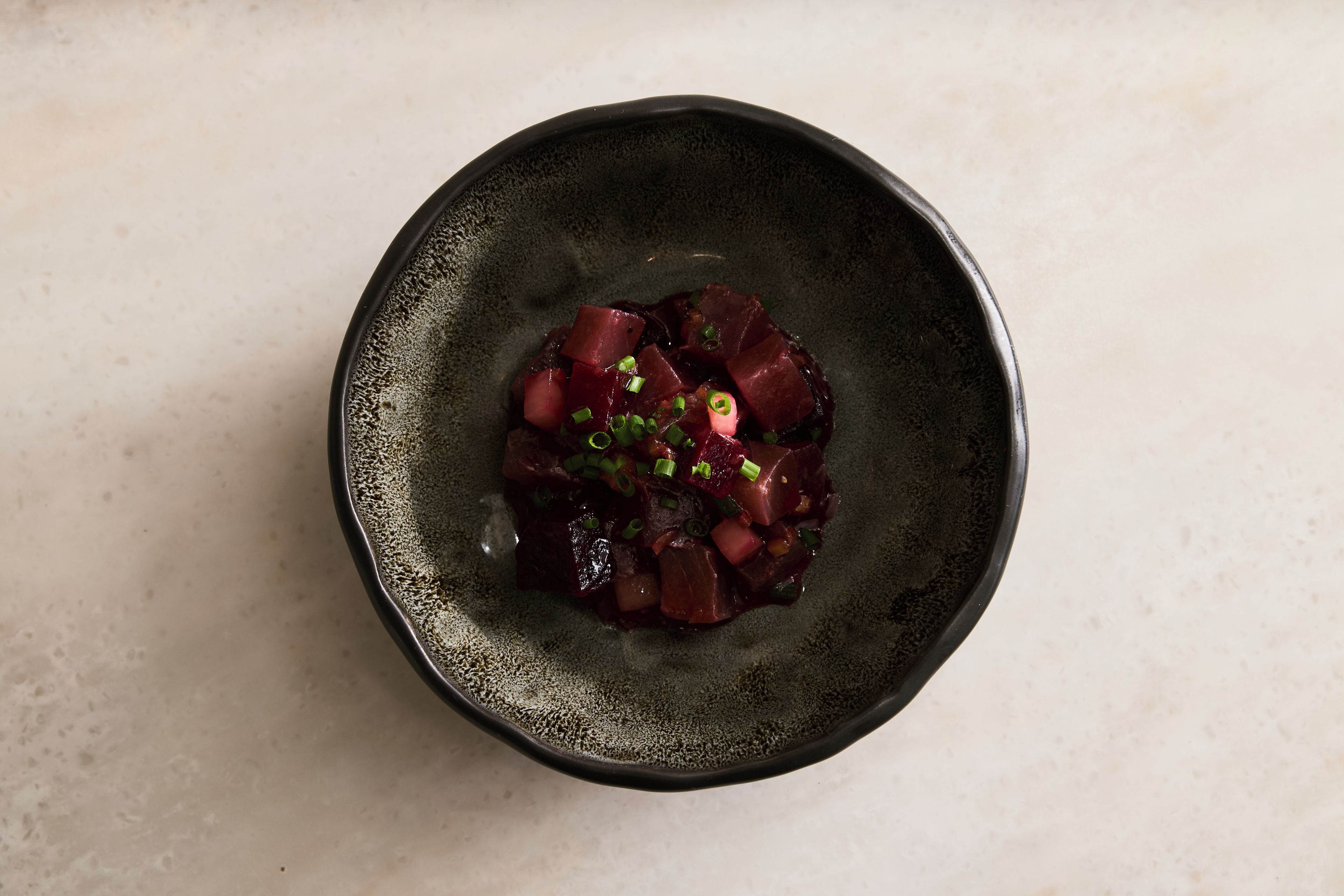 A dark ceramic bowl with a textured surface containing a dish of diced red ingredients, possibly beetroot, garnished with green herbs.