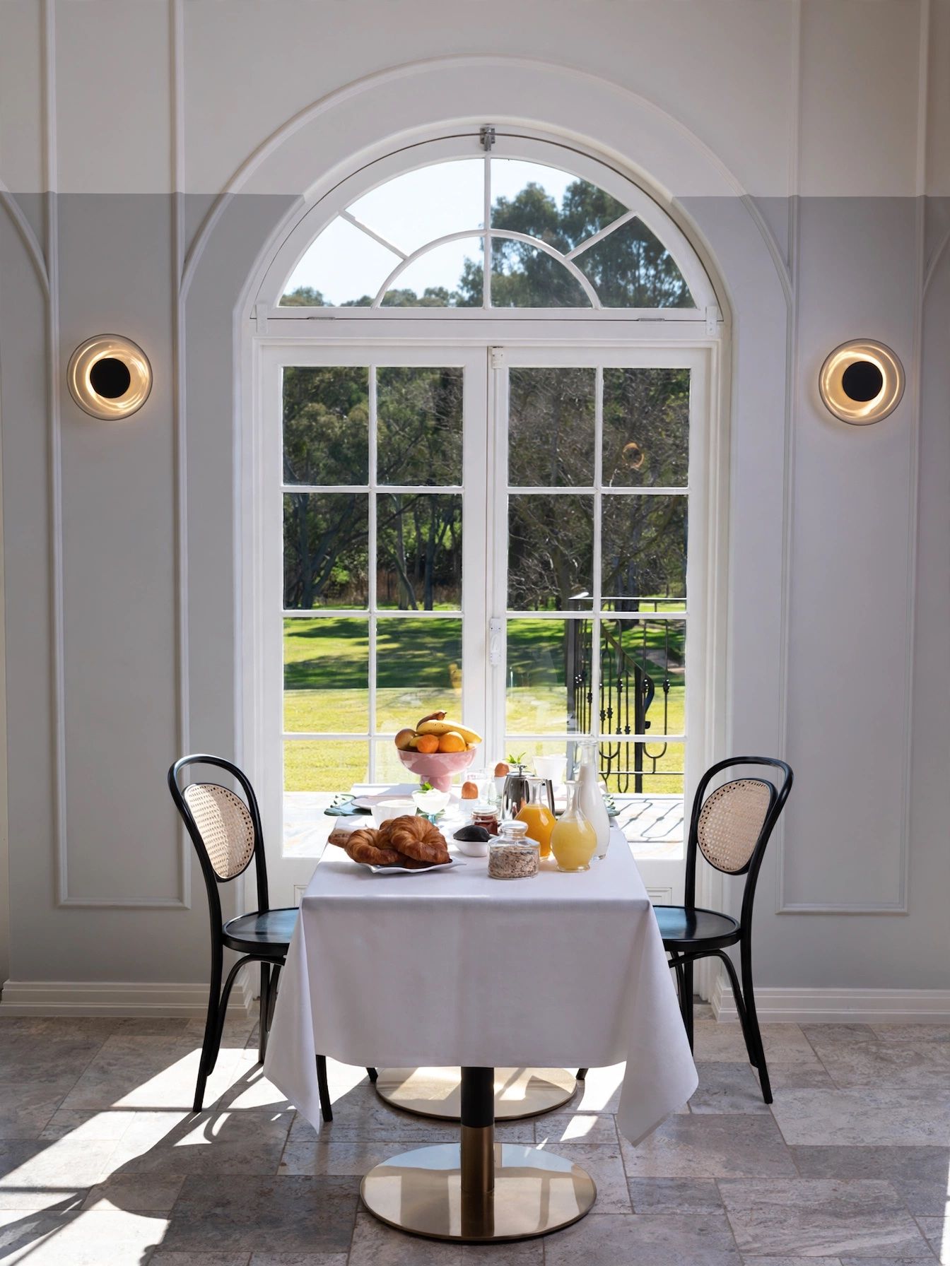 Un comedor con una mesa dispuesta para el desayuno, dos sillas y un amplio ventanal de arco con vistas a un paisaje verde.