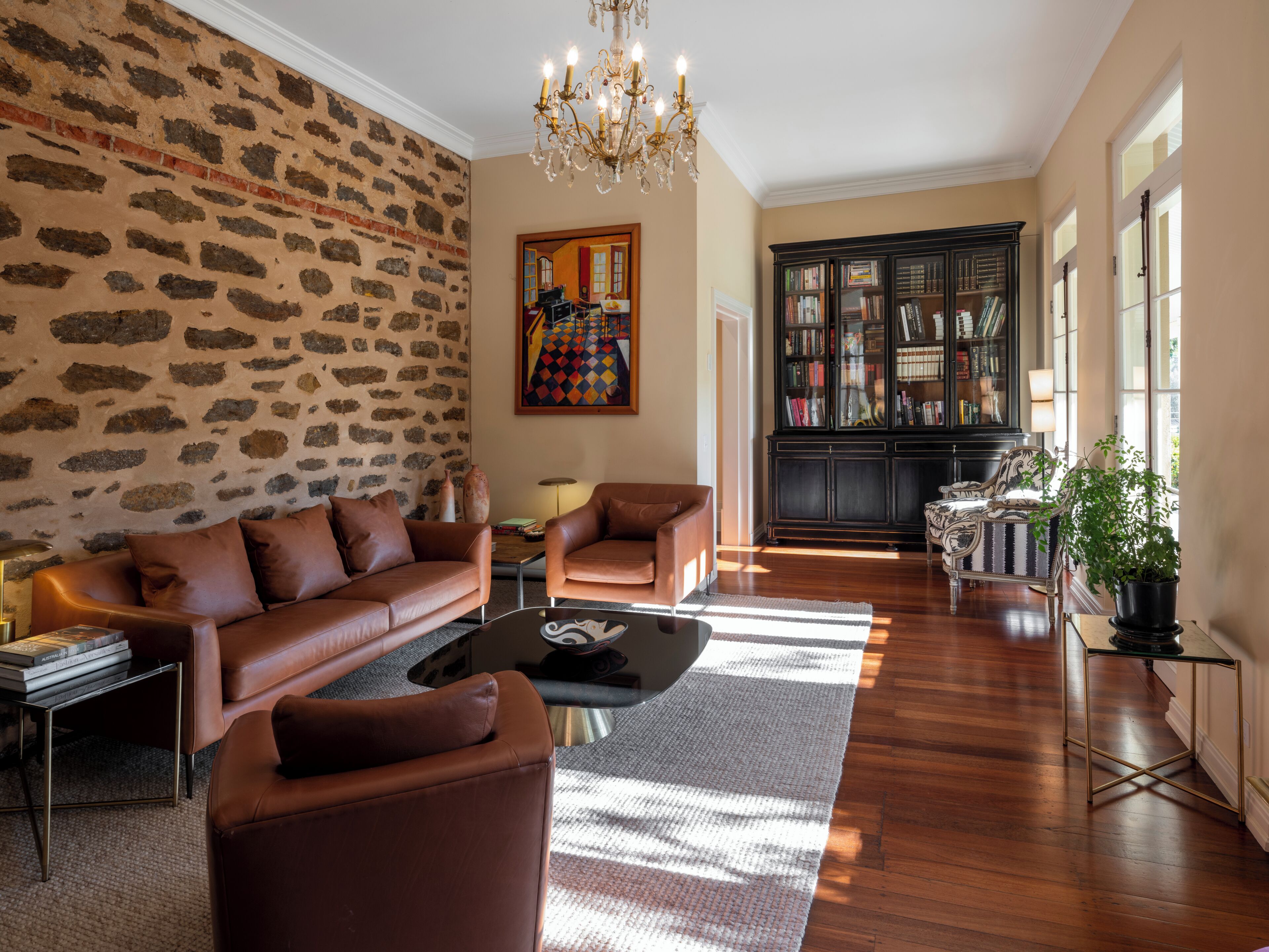 Una sala de estar con pared de piedra, sofás de cuero, una lámpara de araña, una estantería y un ventanal grande.