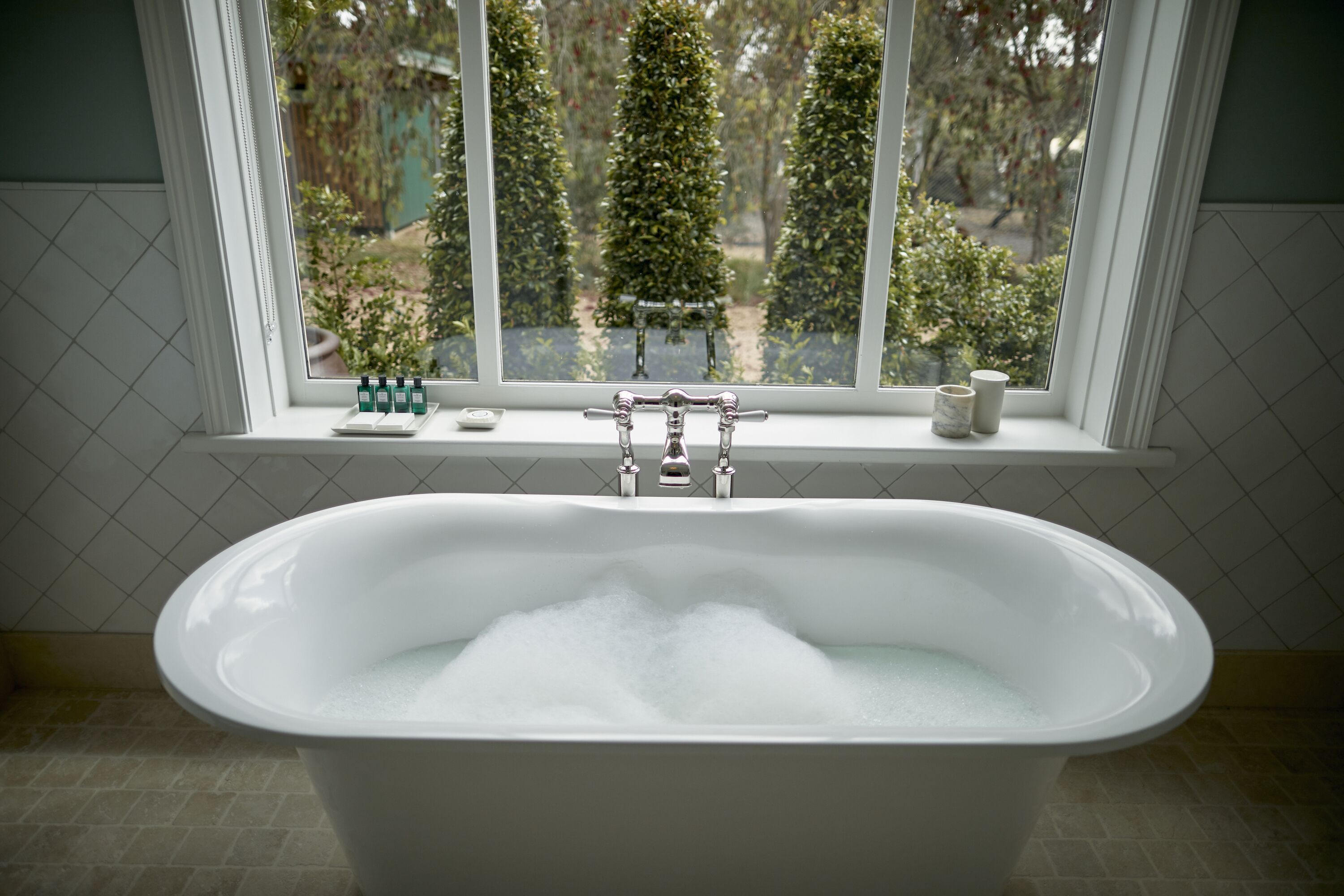 A bathroom with a bathtub filled with bubbles, a window with a view of trees, and toiletries on the windowsill.