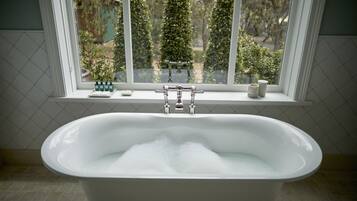 A bathroom with a bathtub filled with bubbles, a window with a view of trees, and toiletries on the windowsill.