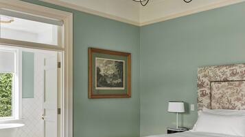 A bedroom with a large bed, a bedside table, a chair, a framed picture on the wall, and a door leading to a balcony.
